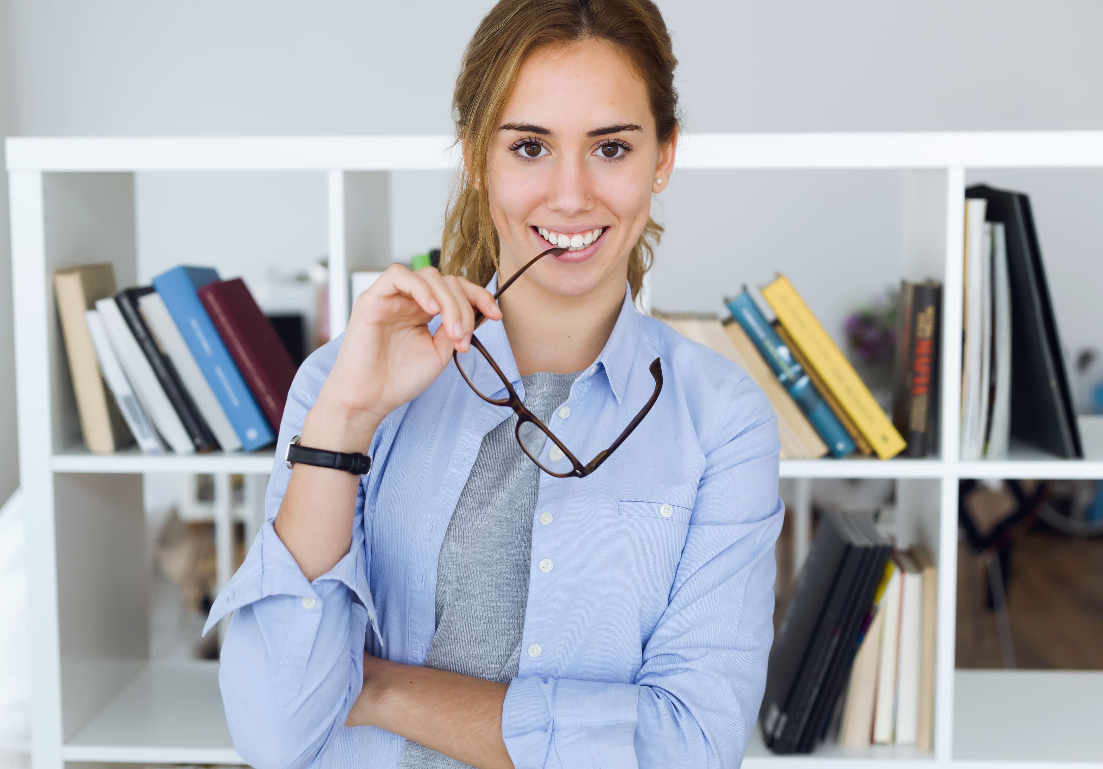 Jeune professionnelle souriante tenant ses lunettes dans un bureau, devant une bibliothèque de travail