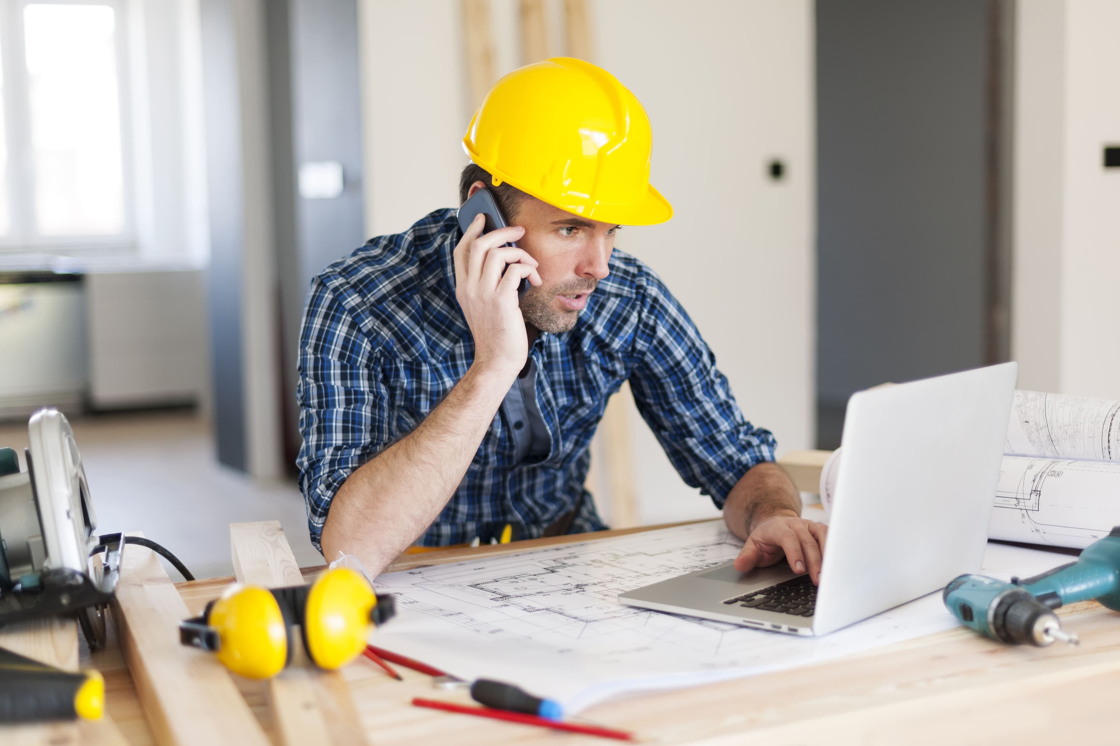 Artisan du bâtiment avec casque de chantier consultant son ordinateur et parlant au téléphone pour gérer un devis de travaux et la TVA Artisan du bâtiment avec casque de chantier consultant son ordinateur et parlant au téléphone pour gérer un devis de travaux et la TVA